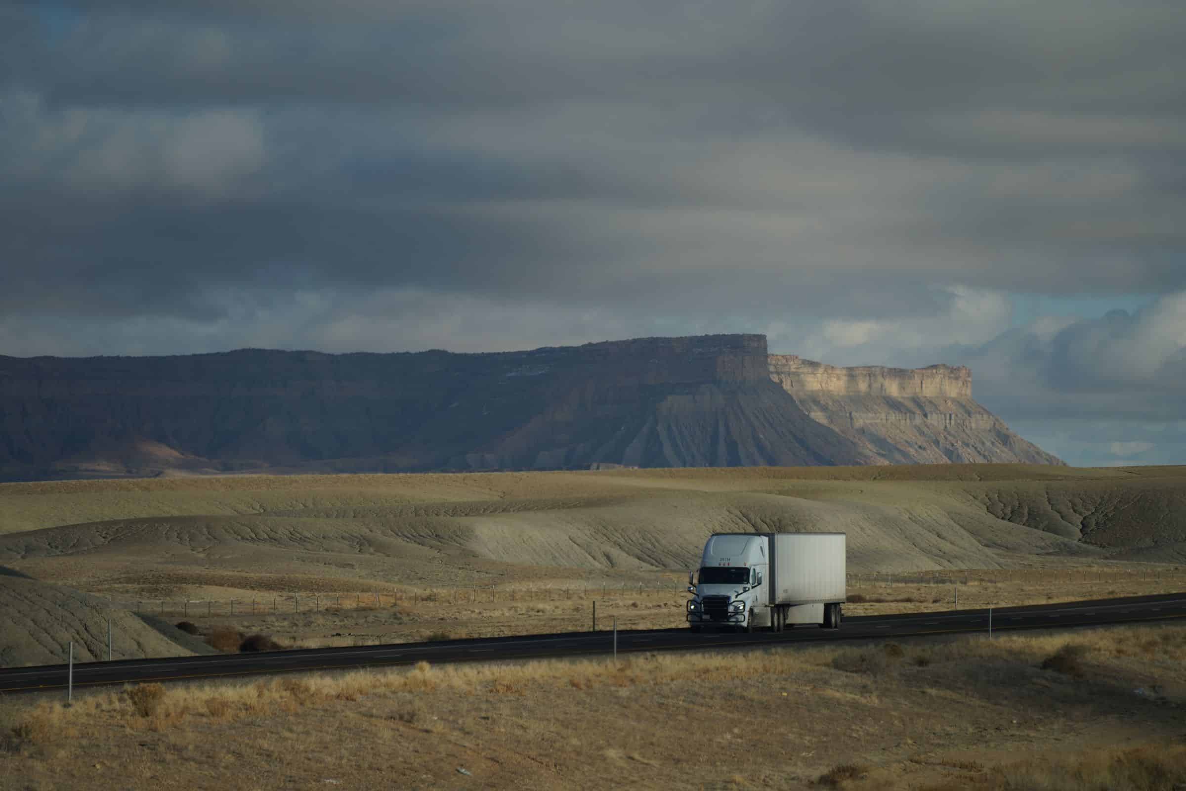 A freight truck driving down a desert highway beneath a dramatic cloudy sky in the American Southwest.